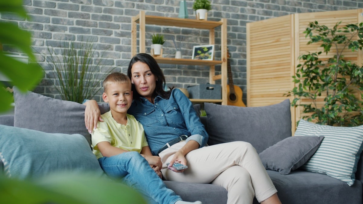 Happy child is watching TV with loving mother at home on sofa holding remote control hugging and laughing. Happiness, family and modern technology concept.